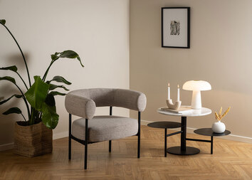 Oakfield Loungestol i stoffet Monza Beige i en stue. Et sidebord dekoreret med en lysestage, en tænd bordlampe og en vase står ved siden af stolen.