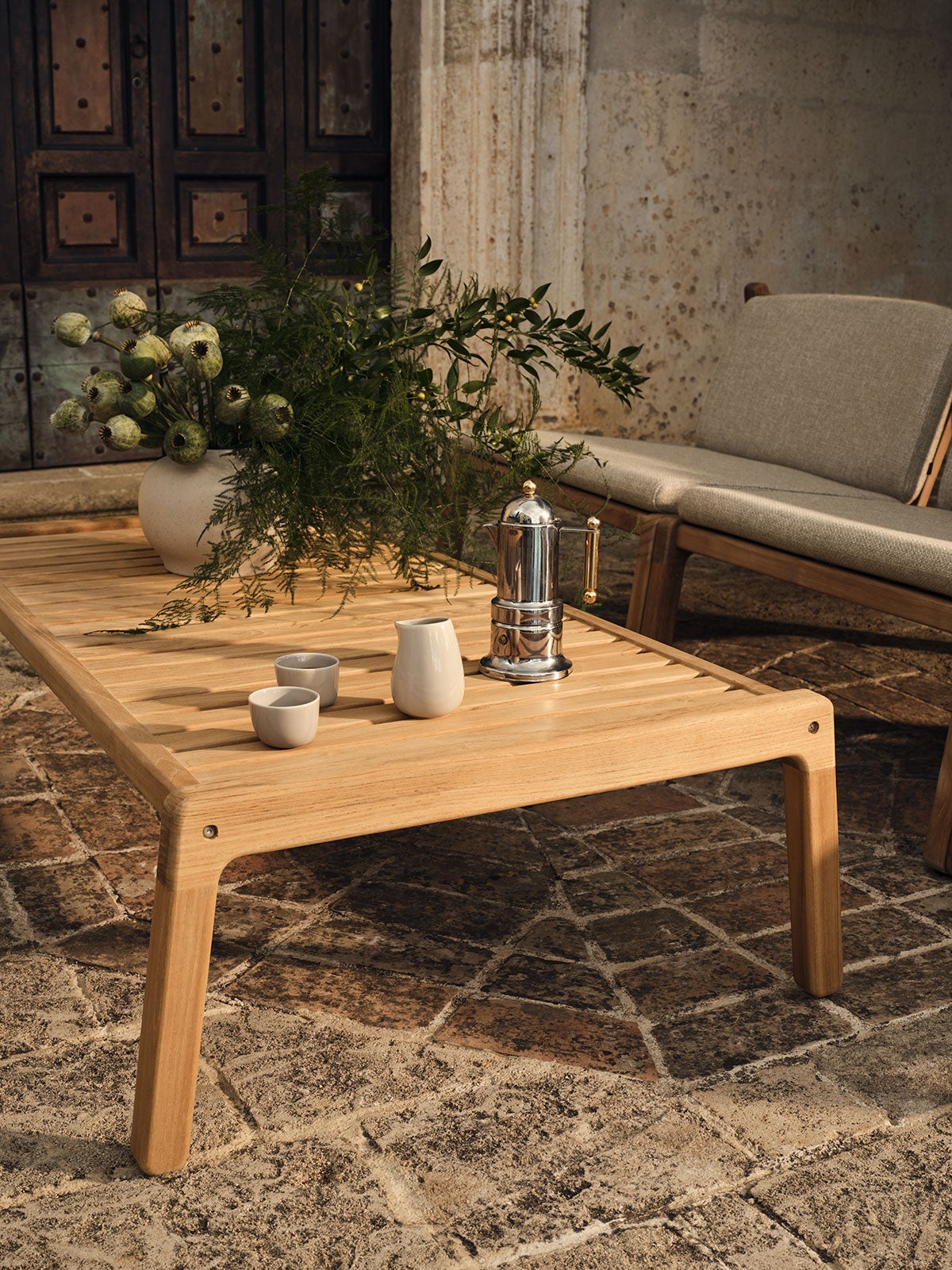 Patio sofabord 120x60 på en solrig terrasse. Sofabordet er dækket med kaffe, kaffekopper og en vase med blomster.