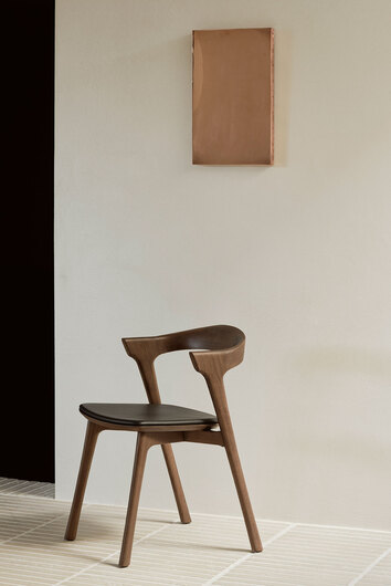 Bok Stol i brunlakeret teak med sort lædersæde, der står op ad en hvid væg med et billede i messing