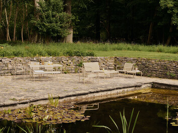 Loungeserien Dapple i farven Cashmere, som står på en stenbelagt udeplads ved siden af en dam