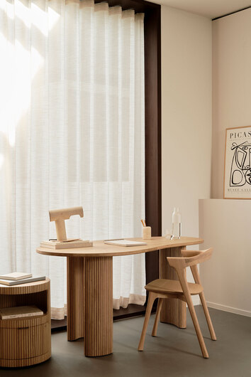 Boomerang Skrivebord i lakeret eg med en bordlampe, en notesbog og en penneholder foran et vindue med lette gardiner