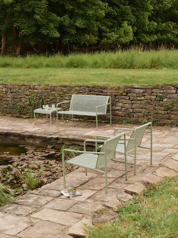 Dapple Loungesofa og Dapple Loungebord 57x57 i farven Tea Green, som står ved siden af en dam. På den anden side af dammen står to Dapple Loungestole i samme farve.