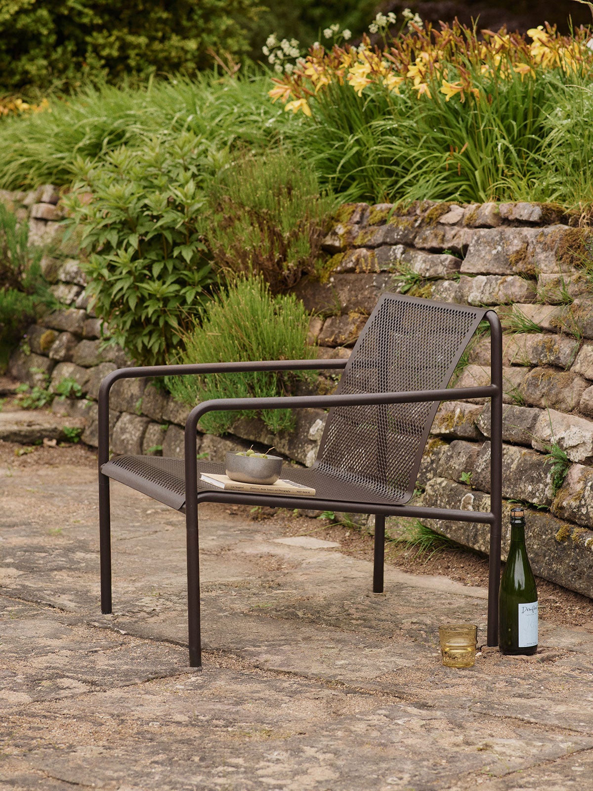 Dapple Loungestol i farven Dark Chocolate, som står på en stenbelagt udeplads fyldt med blomster og grønt