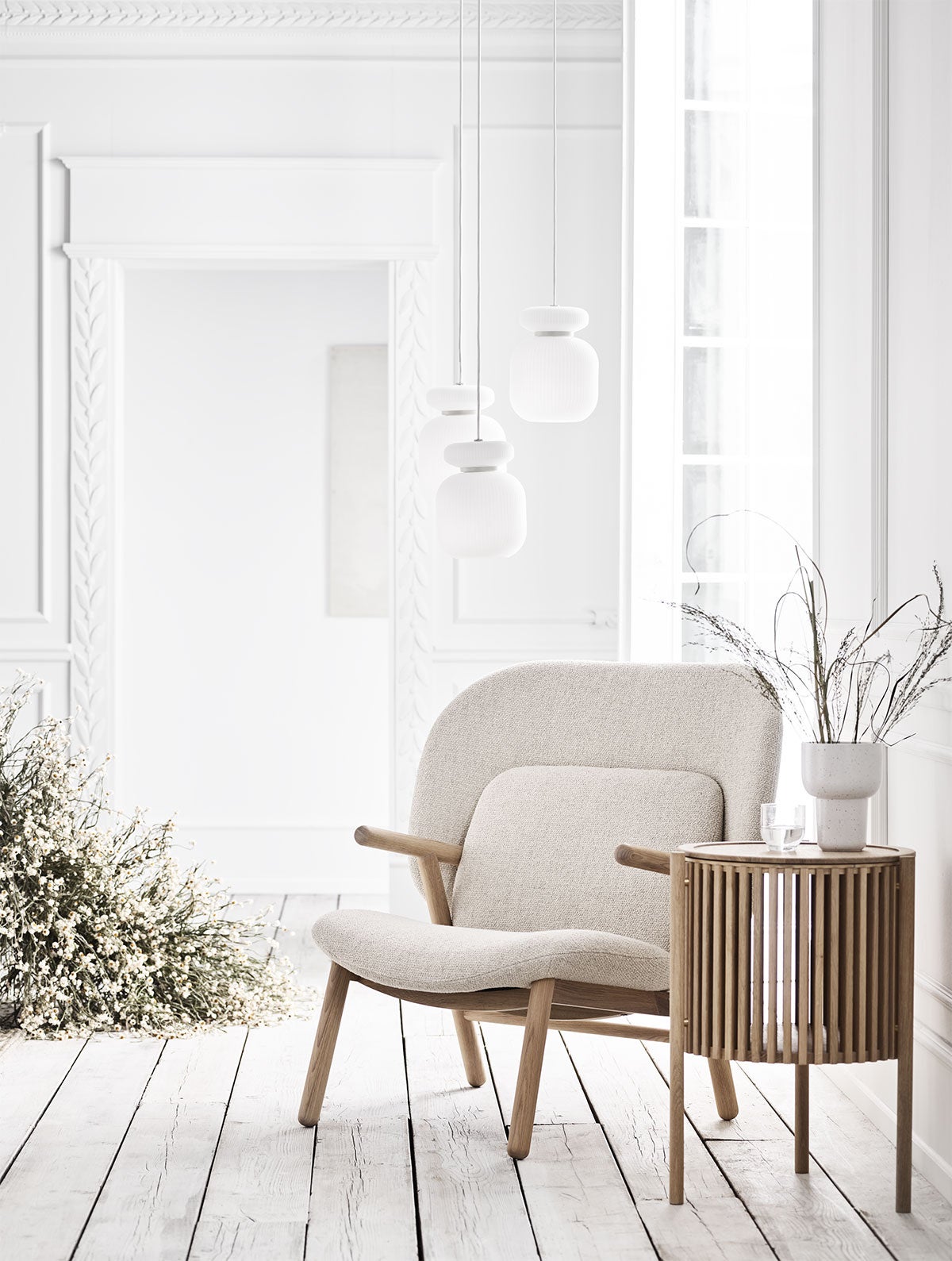 Cosh lænestol betrukket med et lyst stof og stel i olieret eg. Lænestolen står ved siden af et sidebord, som er dekoreret med en vase.