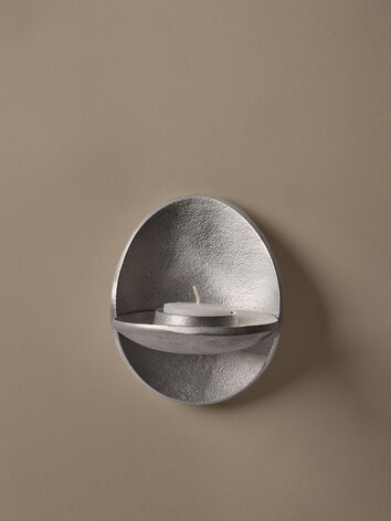 Agapé Vægljusholder i aluminium med et fyrfadslys, som hænger på en beige væg